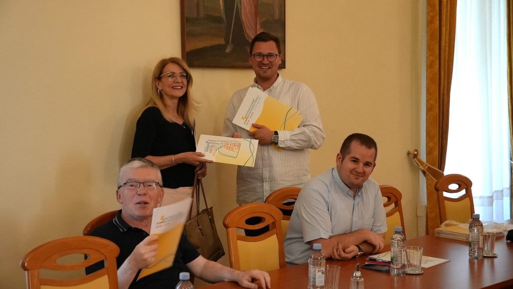 Two people stand by a wall exchanging a certificate while two colleagues sit at a table with documents and water bottles nearby.