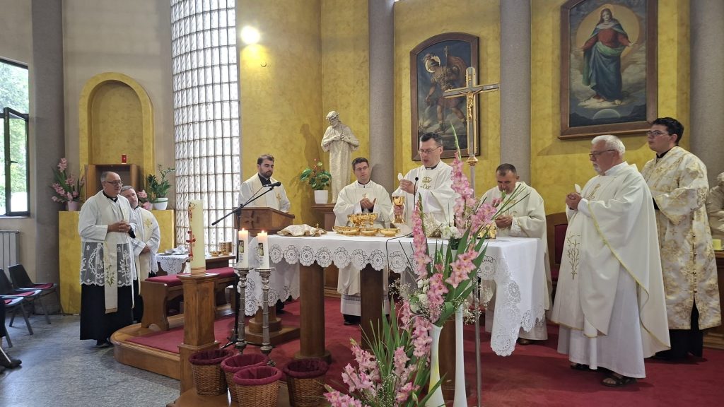 Catholic priests at an altar during Mass, with gold chalices, candles, and pink gladiolus in the foreground inside a church.