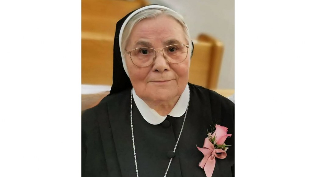 Elderly nun in black habit with white collar, wearing glasses, a pink rose corsage, and a silver chain, facing the camera