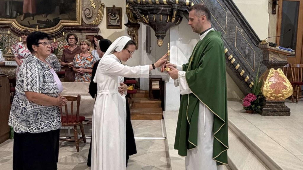Nun in a white habit receives a liturgical item from a priest in green vestments during a church ceremony, with onlookers in the background.