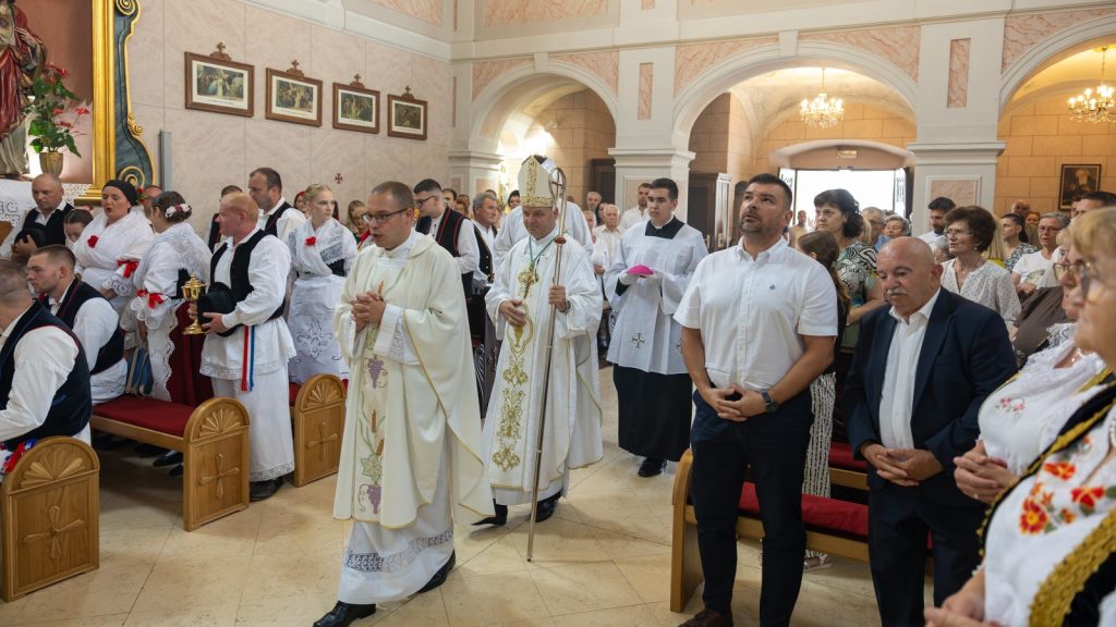 Congregation standing in a church during a ceremony; clergy in white vestments, a bishop with a crozier, and attendees in traditional embroidered clothing.
