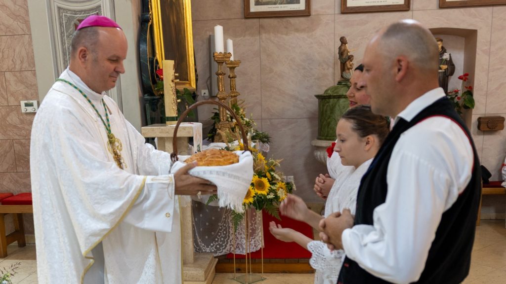 Catholic priest in white vestments hands a loaf of bread on a white cloth to a girl and a man in a church setting with an altar behind them.