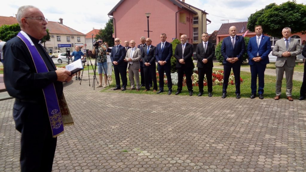 Priest in a purple stole addresses a line of suited men on a cobblestone square during an outdoor ceremony, with photographers in the background.