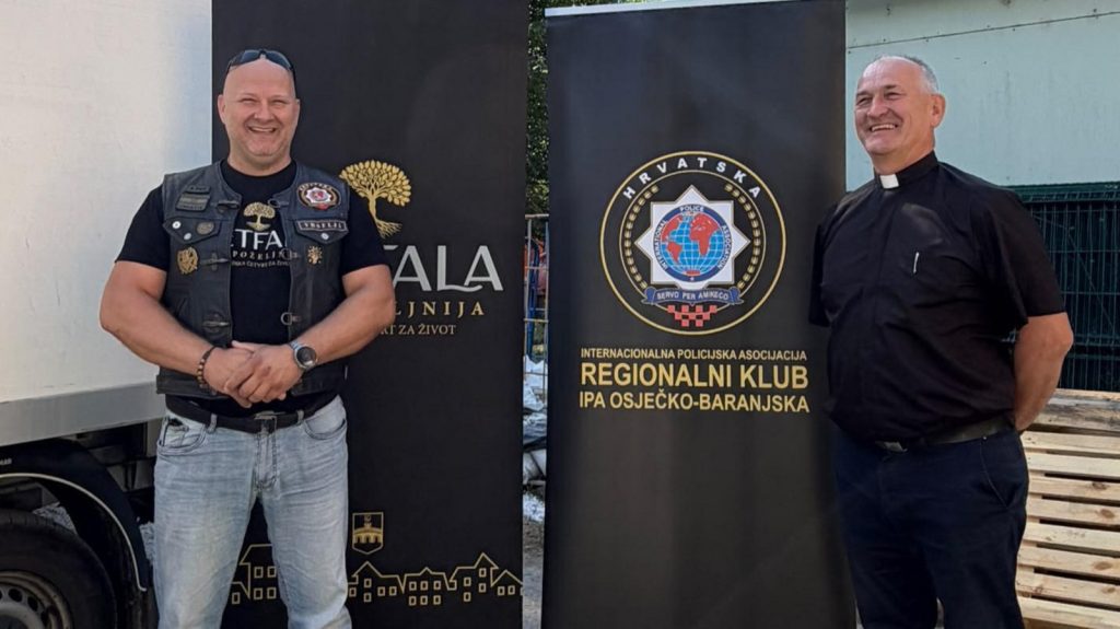 Two men stand beside tall banners; left man in a vest with patches, right man in clerical collar with a smile, outdoors at a club event.