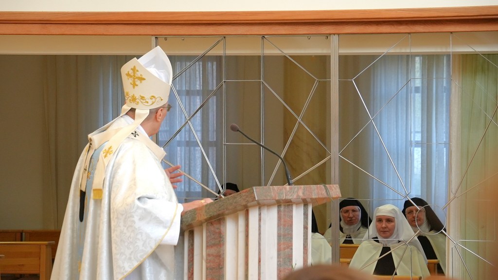 Catholic bishop delivering a sermon at a marble pulpit while nuns sit behind a glass partition in a chapel.