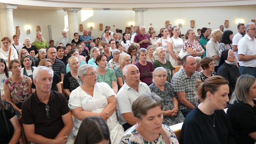 Large crowd of men and women seated in a spacious hall, some standing along the back wall, attentive to an event.