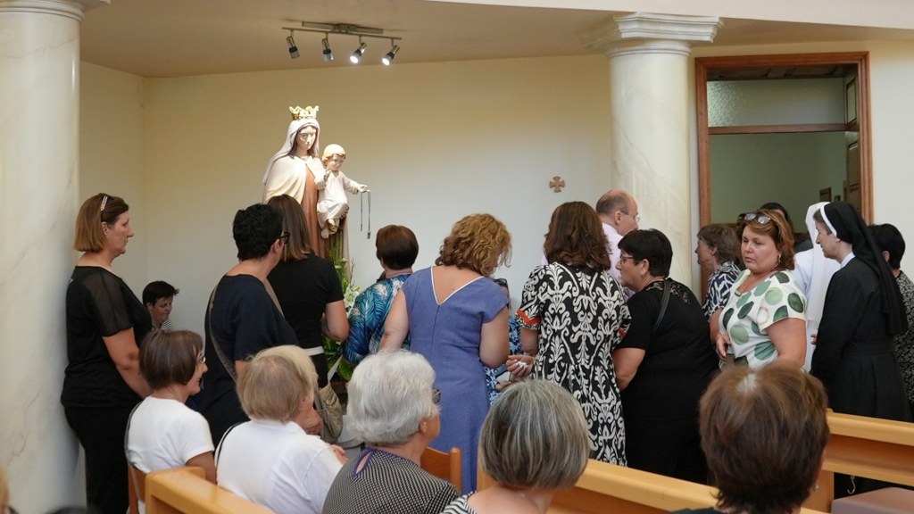 Group of people gathered around a statue of Mary and child in a small chapel with pillars and benches nearby.