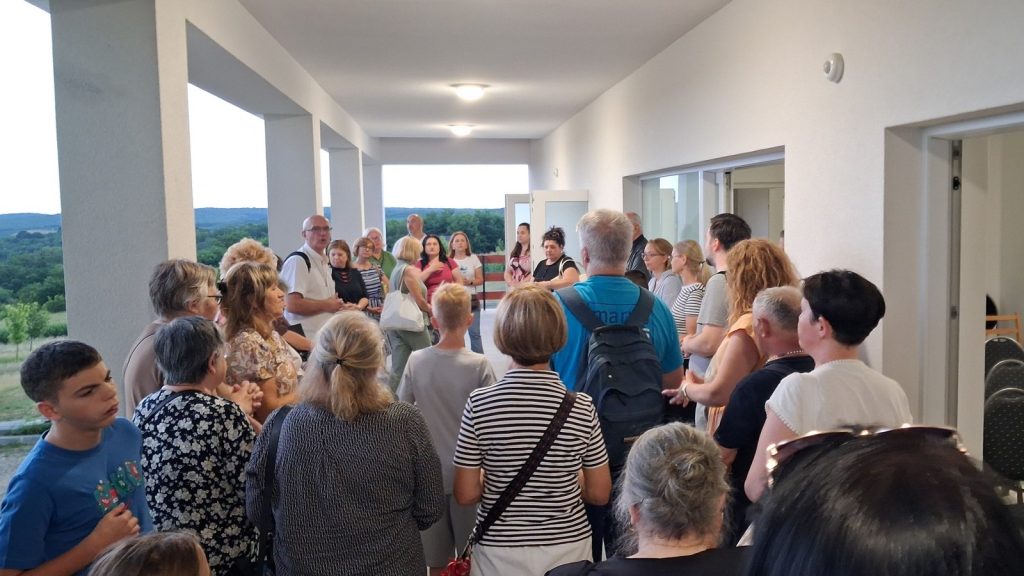 Crowd of adults and children gathered in a bright hallway, listening to a speaker at the far end, with a countryside view outside.