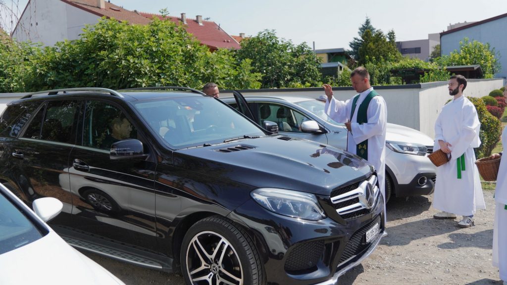 Priests in white robes bless a black SUV with raised hand beside a white car in a parking area. greenery and buildings in the background.