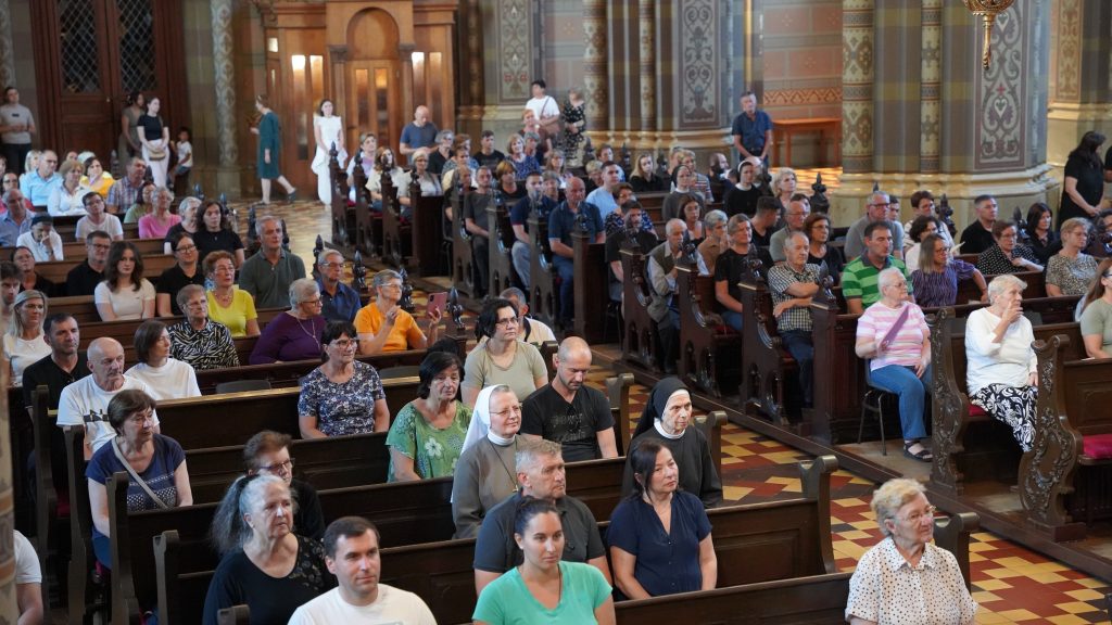 Inside a ornate church filled with people seated in wooden pews, listening to a service amidst decorative columns and tiled floor.