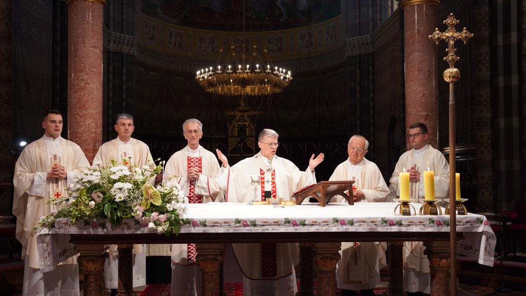 Priests in white robes perform mass at an elaborately decorated altar with a floral arrangement and candles inside a church.