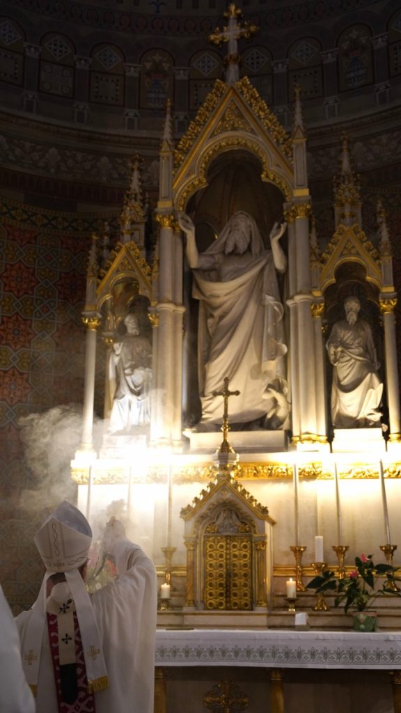 Catholic altar with gold-trimmed statues and central shrine; a bishop in white vestments incenses the area as religious ceremony unfolds, with candles and ornate architecture in the background.