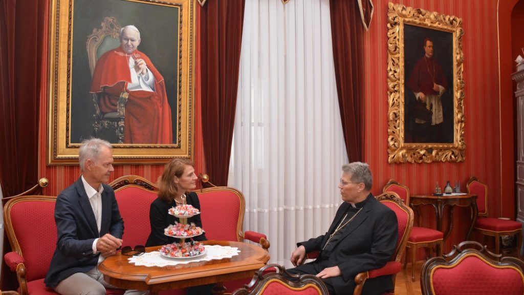Three people sit in a red, ornate room around a table with a candy tower, engaged in conversation.