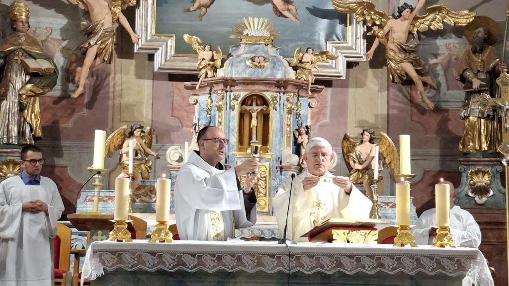 Two priests celebrate Mass at an ornate altar with gold statues, angels, and tall candles in a church setting.