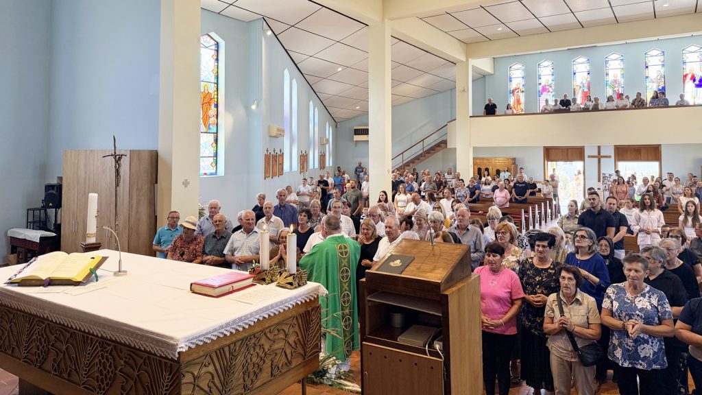 Crowded church during service: priest in green vestment at the altar with candles and an open Bible, congregation standing and praying; stained glass windows in the background.