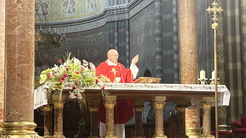 Catholic priest in red vestments raises hands at an ornate altar with a floral arrangement.