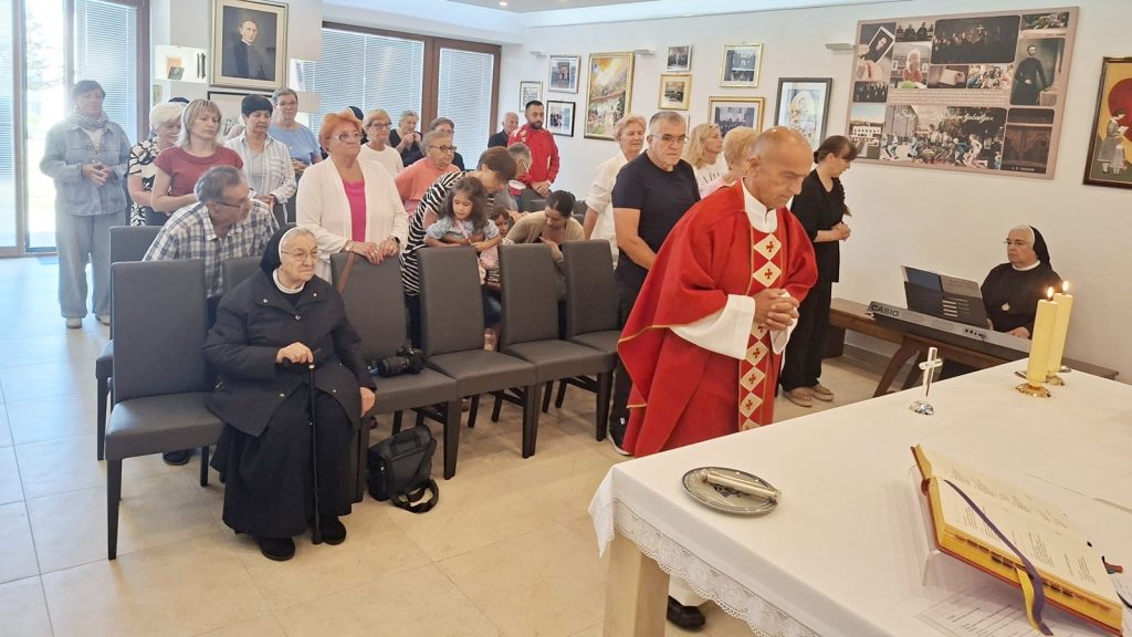 Priest in red liturgical vestments leads a service at the altar as a nun plays a keyboard; an attentive group of elderly parishioners stands and sits in a bright room with framed pictures on the walls.