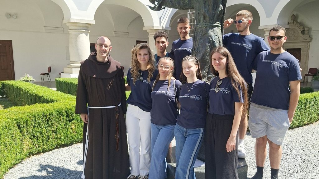 A monk in brown robes with a group of students in navy shirts posing outdoors in a bright courtyard with arches and a statue behind them.