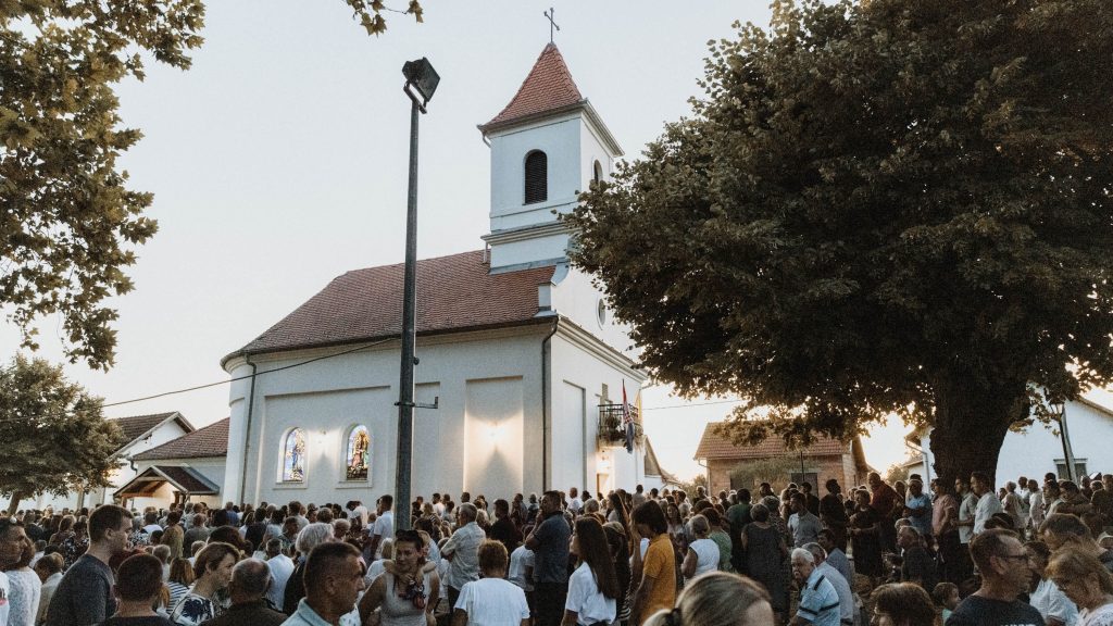 Crowd of people gathered outside a white church with a red-tiled roof and small steeple at dusk, framed by trees and a street lamp.