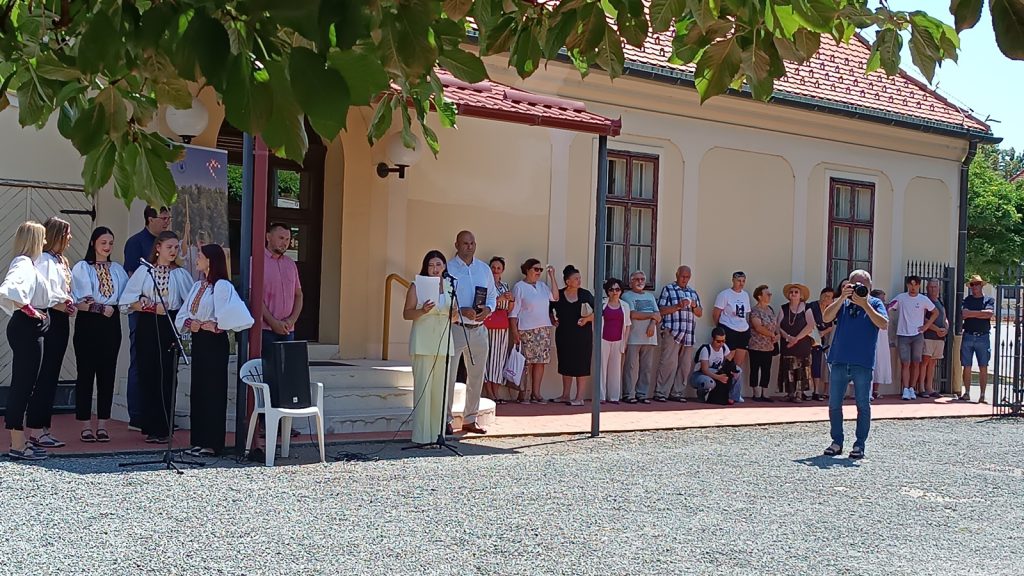 Outdoor performance with young women in traditional white blouses singing on a small stage beside a pale building; a crowd watches.