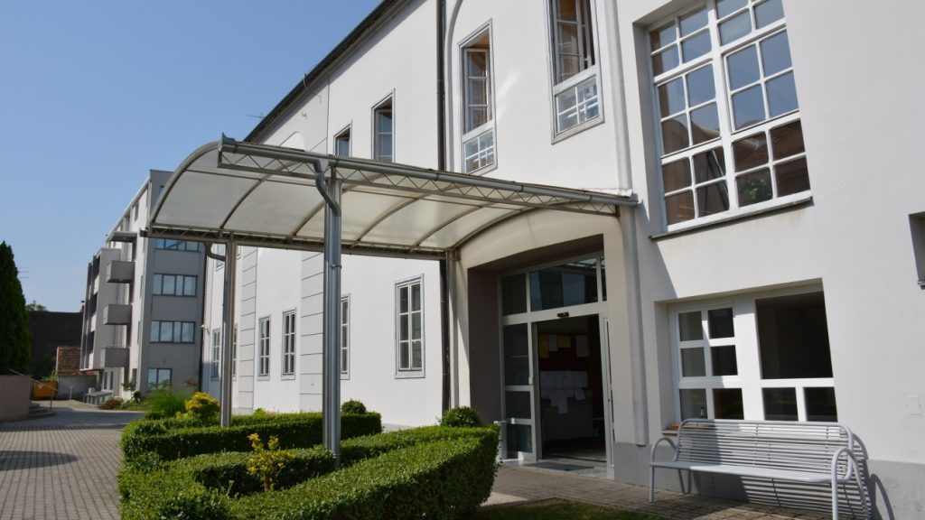 Entrance of a white campus-style building with a curved translucent canopy over glass doors and trimmed hedges nearby, plus a bench nearby.