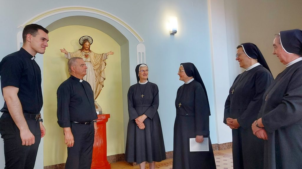 Five clergy members stand in a light blue hallway, four nuns in black habits and two men, facing a statue of Jesus with outstretched arms.