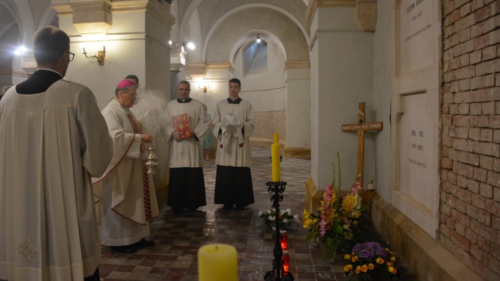 Catholic priests in white robes standing in a church aisle, bishop with censer and book, others holding a white cloth and looking on. A cross, candles, and flowers sit nearby.