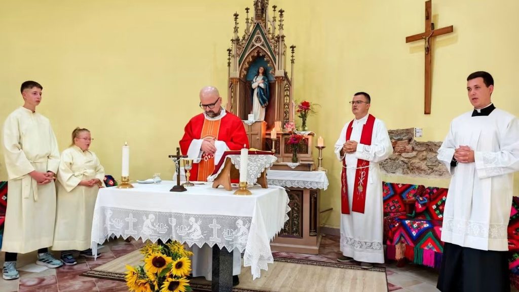 A priest in red vestments leads Mass at an altar inside a church, flanked by two clergy and two young altar servers wearing white robes.