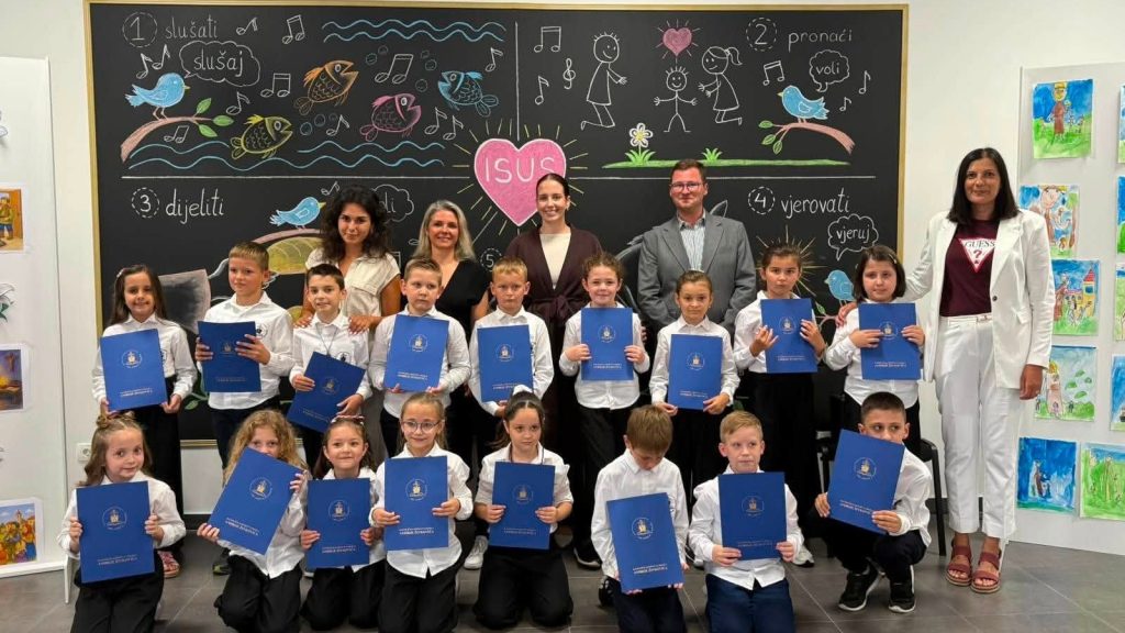 Group of children in white shirts hold blue folders during a certificate ceremony, with teachers behind them in a classroom.
