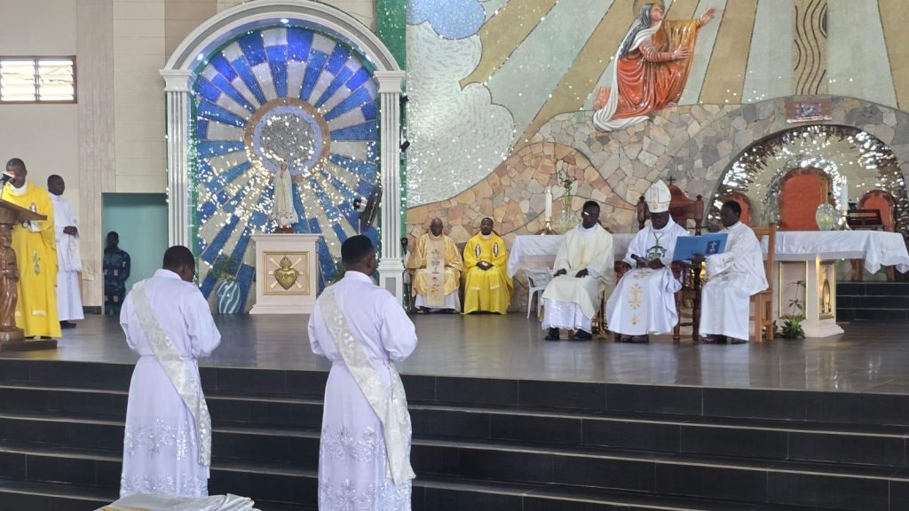 Clergy in white robes and yellow vestments standing on a church altar during Mass, with a mosaic backdrop and a statue at the center.