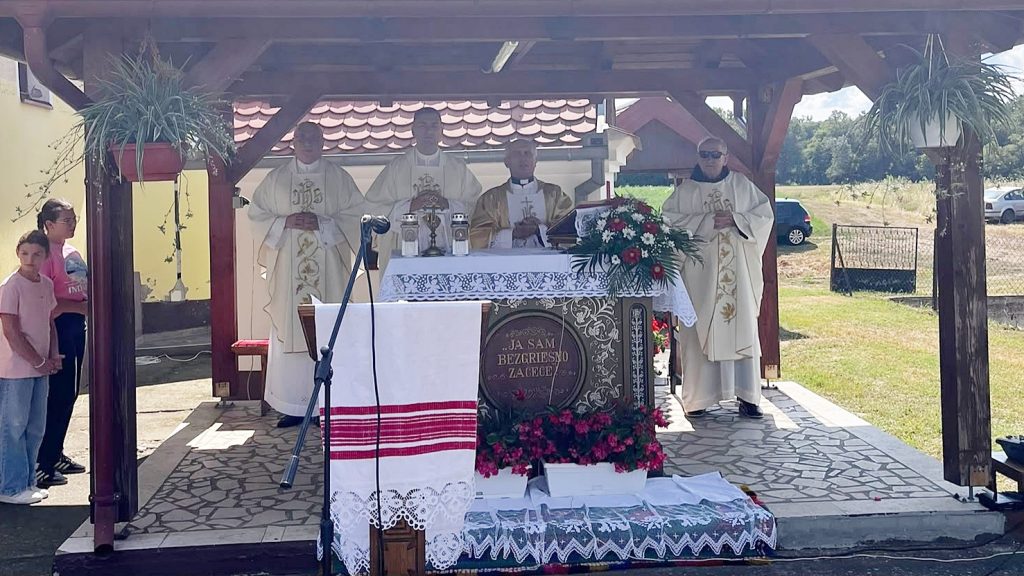 Outdoor Catholic altar under a wooden shelter with four priests in white vestments, floral arrangements, and a microphone stand.
