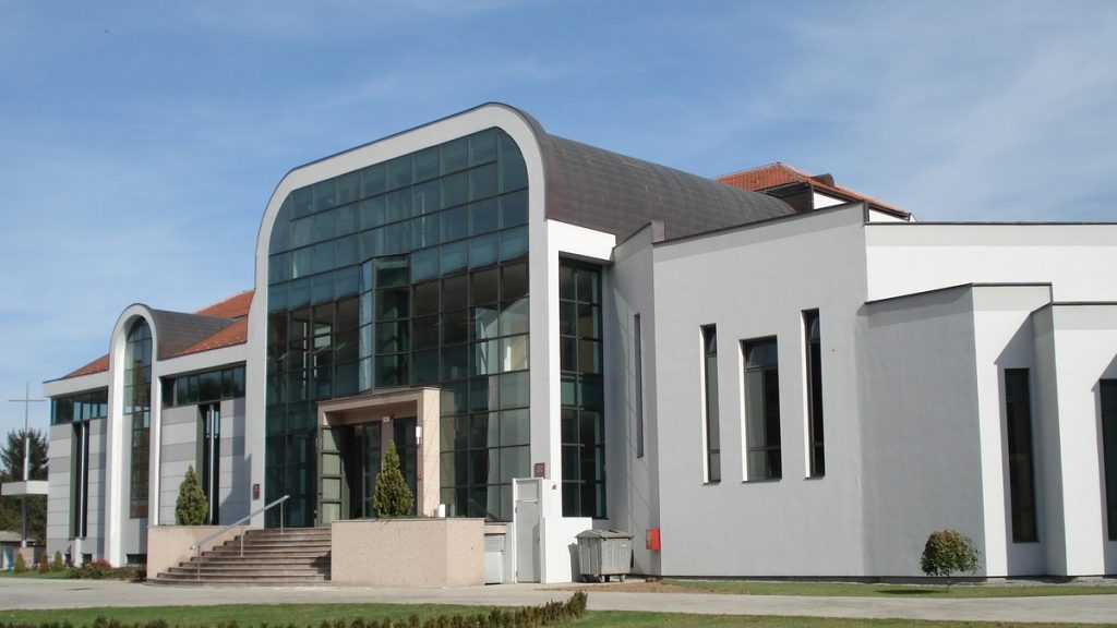 Modern building with a large glass front and curved roof, steps leading to the entrance.