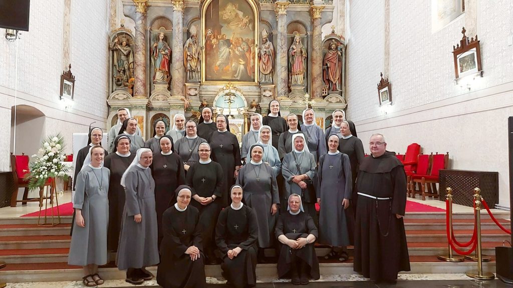 Group of nuns and a priest posing for a photo inside a church with an ornate altar and paintings behind them.