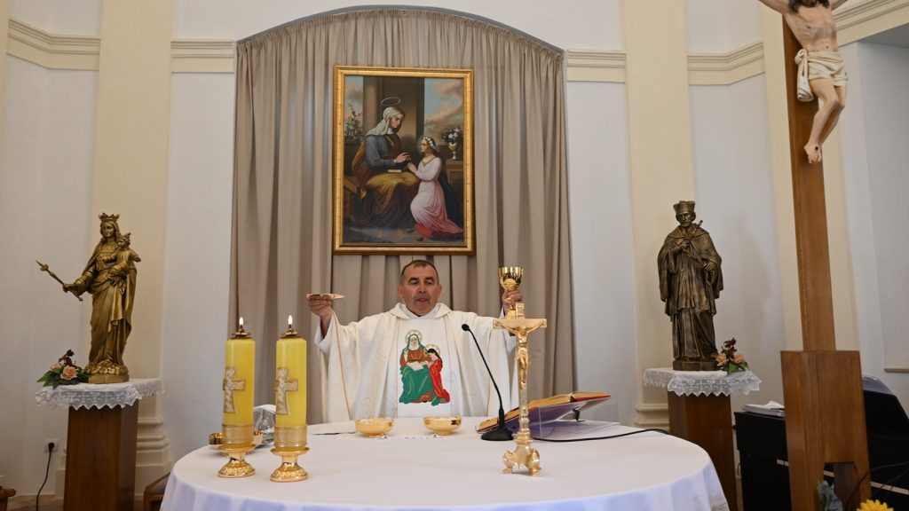 Priest conducting Mass at an altar with a chalice, host, and two burning candles amid statues.