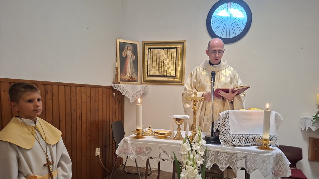 Priest in gold vestments reads from a red book at a lace-draped altar with candles and a crucifix nearby.