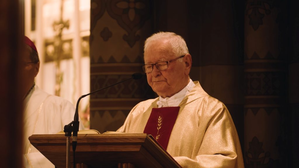 Elderly priest in golden vestments at a wooden lectern reading from a book during a church service, with a microphone nearby.