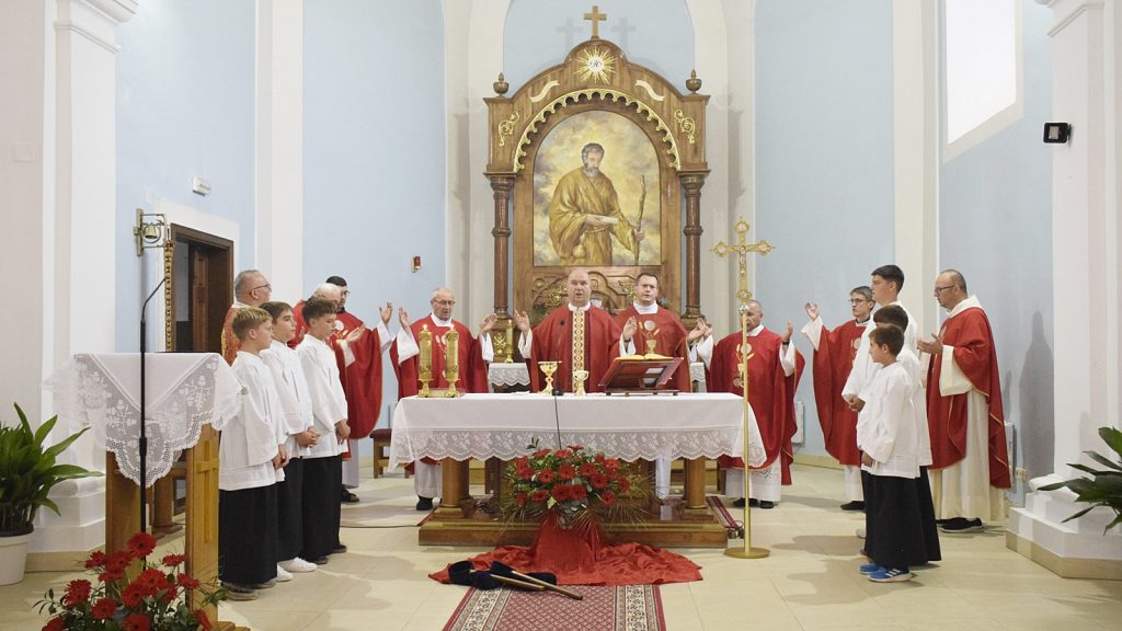 Catholic service at an ornate altar: priests in red vestments lead prayer, flanked by altar servers in white robes.