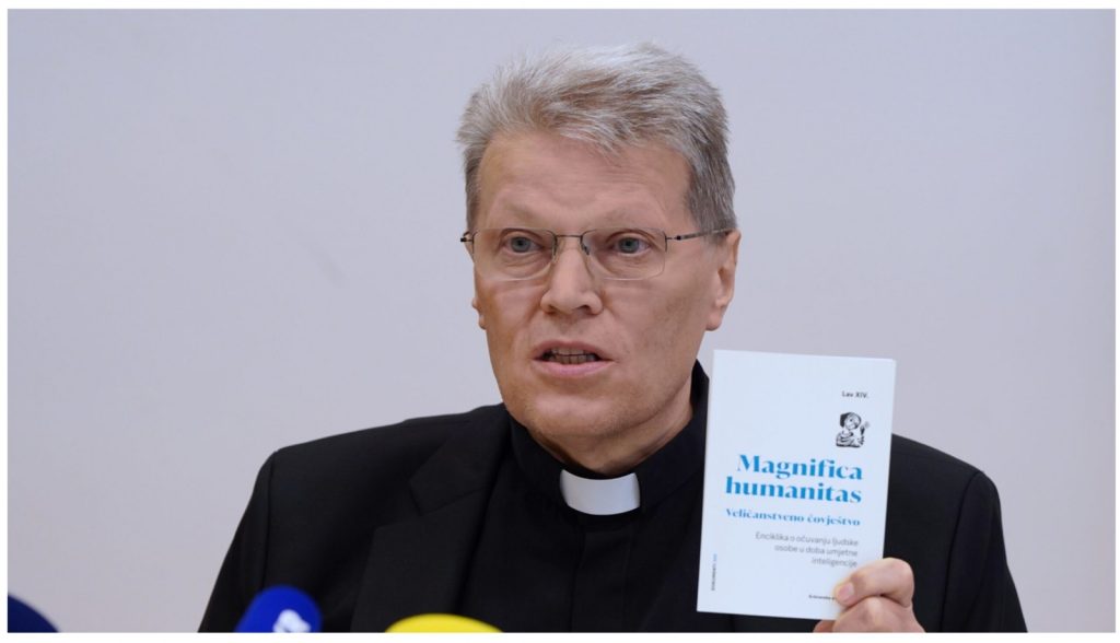 Cleric in black suit holding a white booklet titled 'Magnifica humanitas' with blue text, shown at a press event.