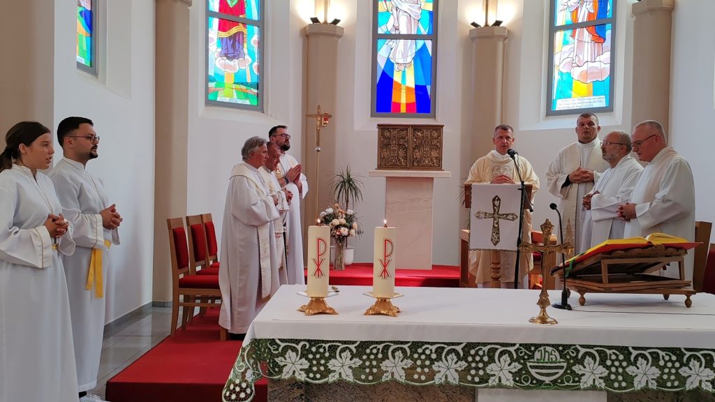 Priests and altar servers stand in a church near the altar during a service, with stained-glass windows behind them and candles on the altar table.