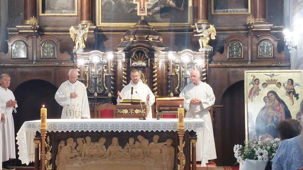 Priest in white vestments speaks at an ornate church altar, flanked by two assistants, with lit candles and intricate woodwork behind.