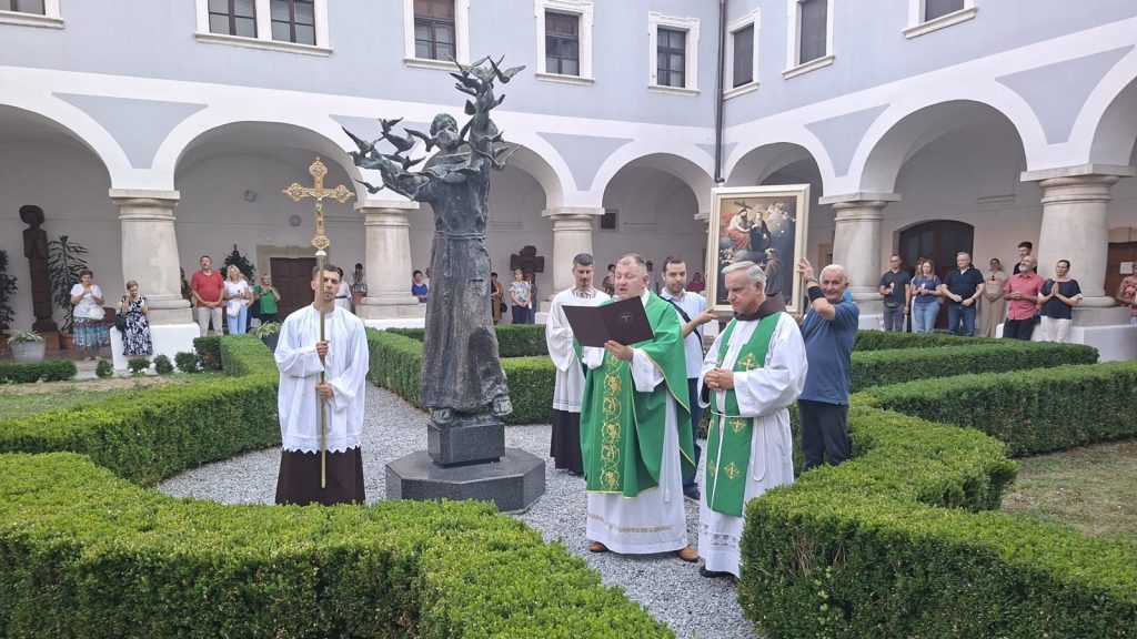 Catholic priests in green vestments conducting a liturgical ceremony around a bronze statue in a cloistered courtyard, as worshippers observe from the arches.