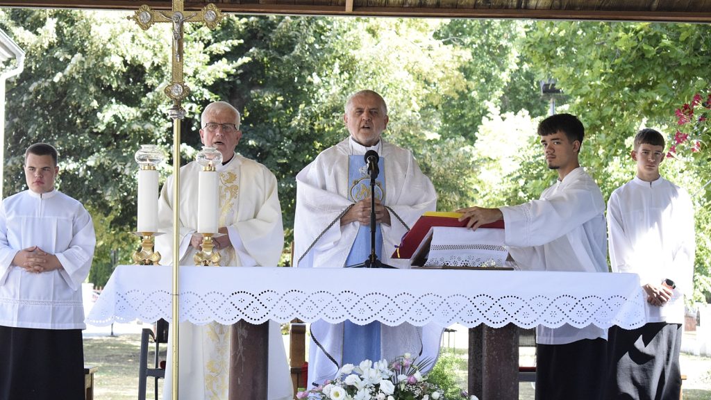 Priest delivering an outdoor Mass at an altar, flanked by two acolytes in white robes beside a crucifix and lit candles.